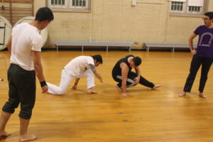 Man watches two others work a modified couch position necessary in capoeira. 